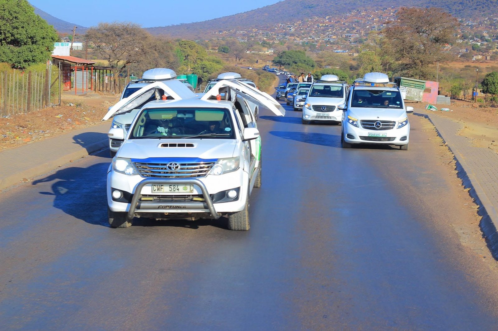 A line of white funeral procession vehicles, including a Toyota Fortuner and Mercedes Vito with roof carriers, travelling along a road past hillside housing